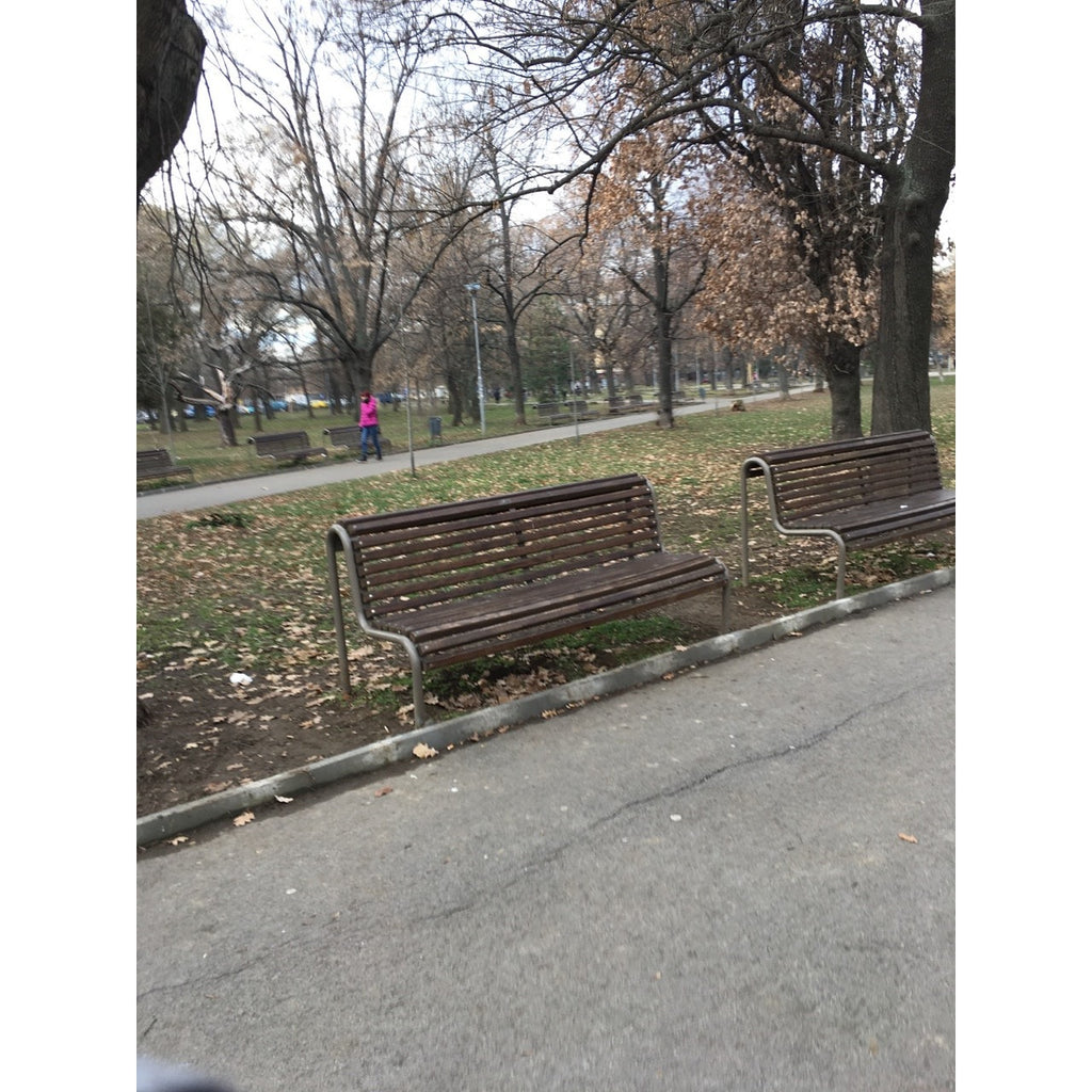 Дървена градинска пейка в парк с паднали листа и пешеходец на заден фон.