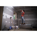 Жена работи с четка на стълба в помещение с гипсокартон и инструменти.