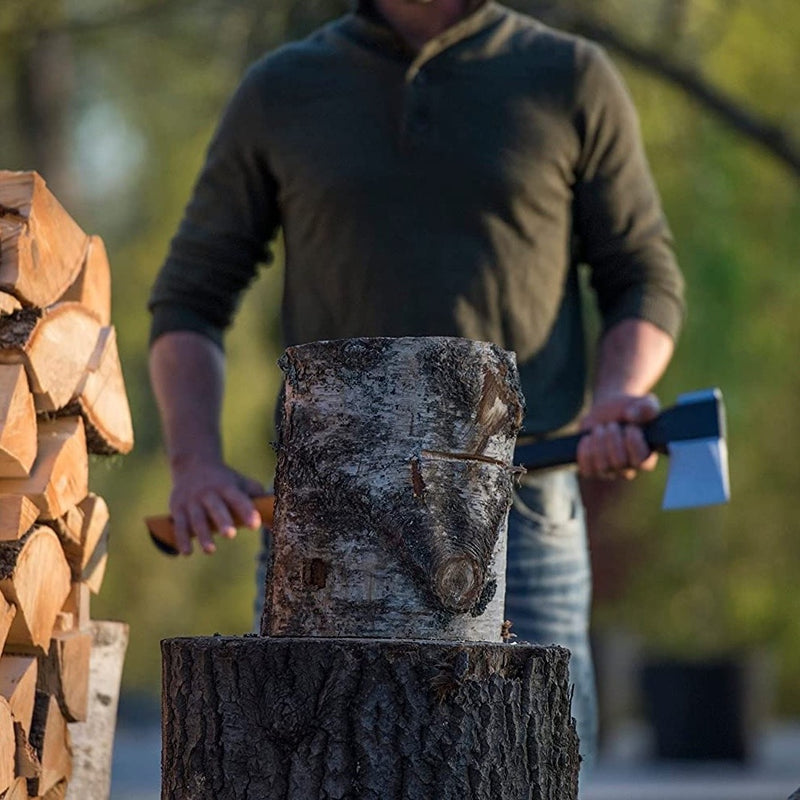 Мъж цепи дърва с брадва Fiskars X27, дървени трупи и дървен блок на задния план.
