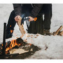 Мъж с брадва Fiskars X7 XS реже дърва за огън в сняг, около лагерен огън.