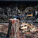 Брадва Fiskars X21 L, зацепена в дърво, на фона на купчини цепени дърва.