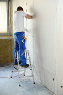 Работник на двустранна алуминиева стълба KRAUSE DOPPLO, извършващ ремонти на стена в интериор.