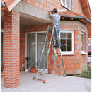 Мъж на двойна алуминиева стълба KRAUSE TRIMATIC при монтаж на къща с червена тухлена фасада.