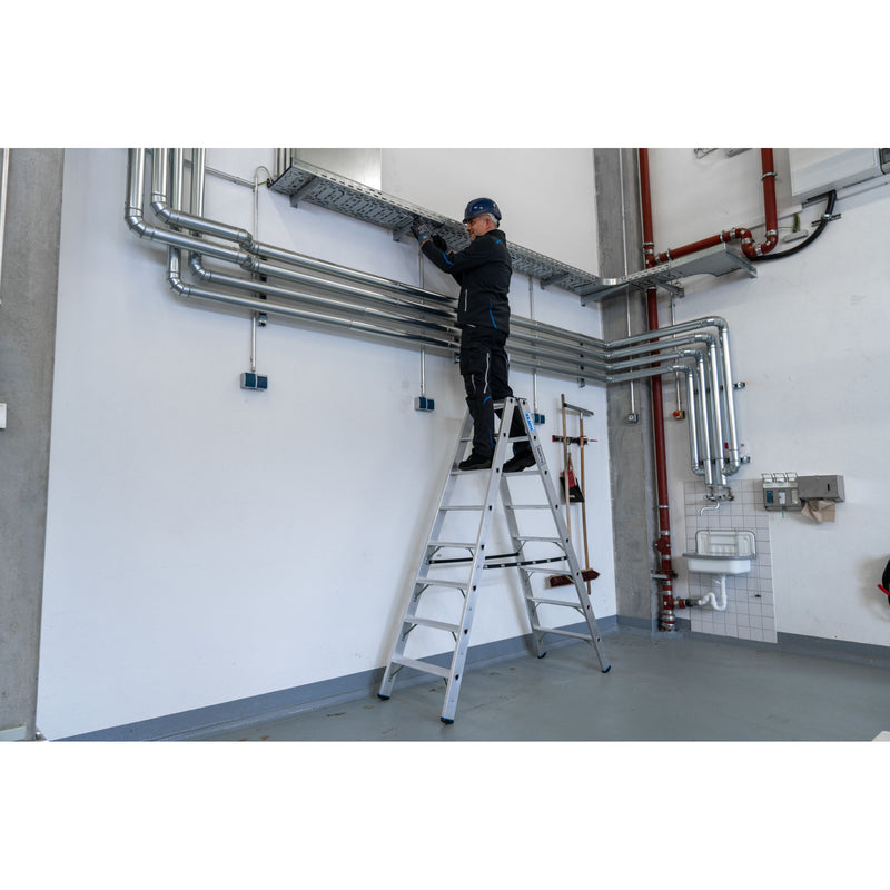 Човек на професионална алуминиева телескопична стълба Krause STABILO, работещ по тръбопровод в индустриална среда.