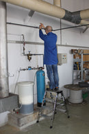 Работник използва алуминиева стълба KRAUSE, за да извърши монтаж на тръби в индустриално помещение.