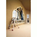 Работник на телескопична дъска Little Giant™ пред голям прозорец, боядисва стена.