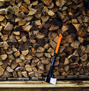 Брадва за цепене Fiskars X17 M, поставена до купчина нарязани дърва на фона.