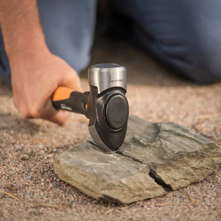 Строителен чук Fiskars IsoCore™ размер M в действие върху камък с ергономична дръжка и система IsoCore™ за намаляване на вибрациите