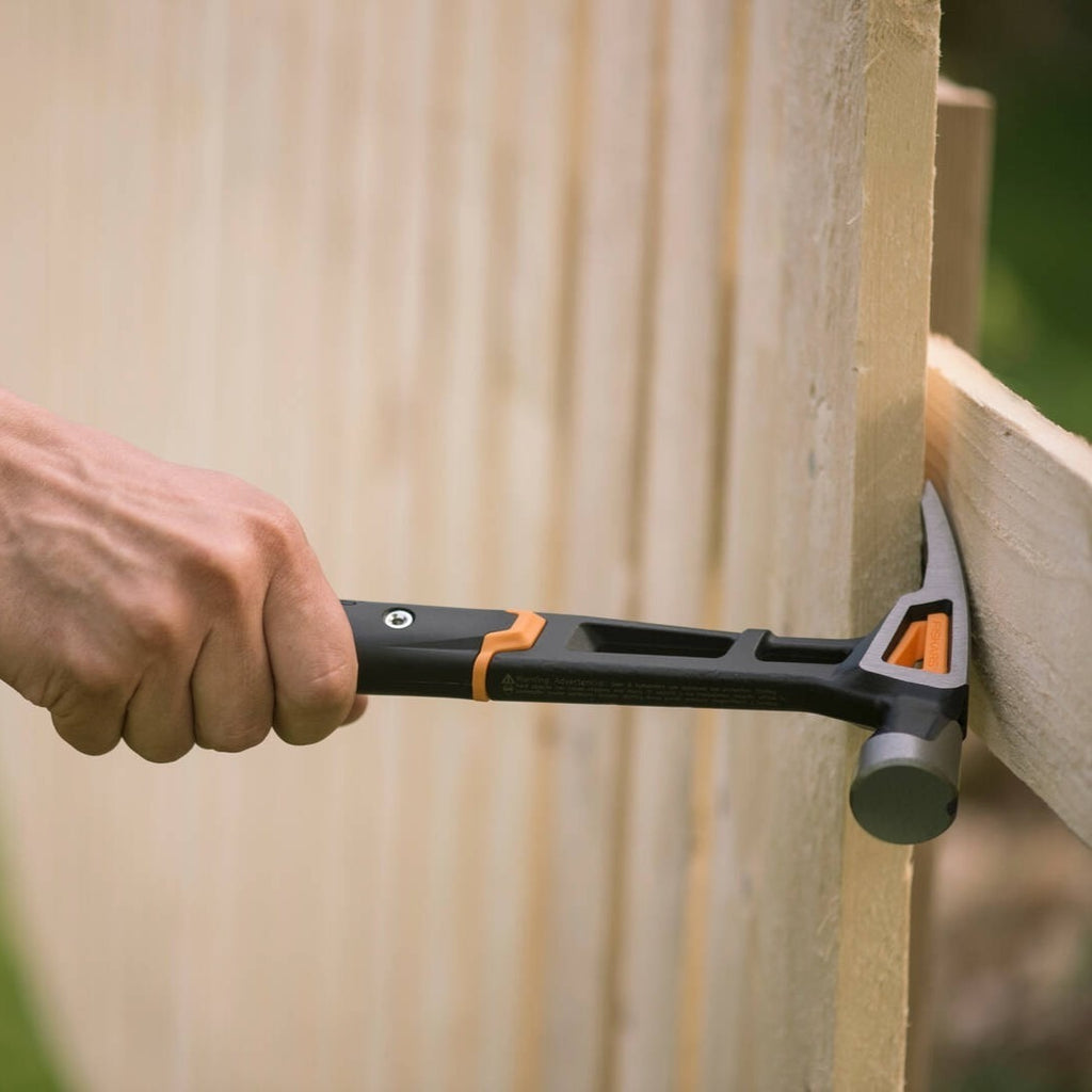 Alt Tag: Универсален чук Fiskars IsoCore™ размер L в действие - поставяне на гвоздей в дървена ограда с ергономична черно-оранжева дръжка