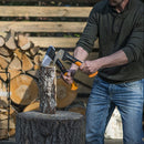 Мъж използва брадва за цепене на дърва X11 на труп в градината, заобиколен от нарязани дърва.