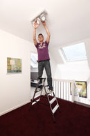 Мъж монтира осветление на стълба KRAUSE DOPPLO в светла стая с високи тавани.