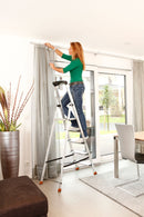 Човек използва алуминиева стълба KRAUSE Sepuro 4+1 за поставяне на перде в домашна обстановка.