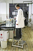 Работник в лаборатория в бяла престилка, стоящ на стълба KRAUSE STABILO, извършващ инженерна работа.