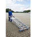 Мъж носи мобилна складова стълба с платформа KRAUSE STABILO, оборудвана с колела за лесно транспортиране.