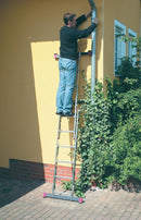 Човек използва алуминиево скеле-стълба KRAUSE за работа на височина пред жълта стена с растения.