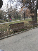 Дървена градинска пейка в парк с паднали листа и пешеходец на заден фон.